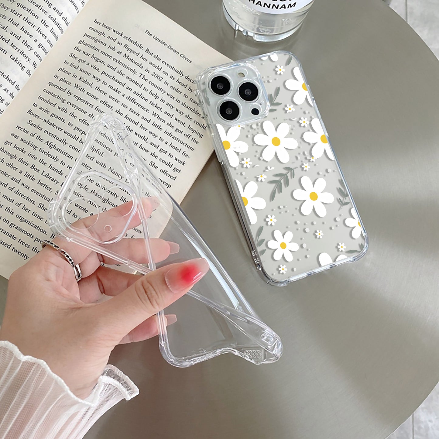 Clear phone case with floral design held by a hand, next to an open book and glass.