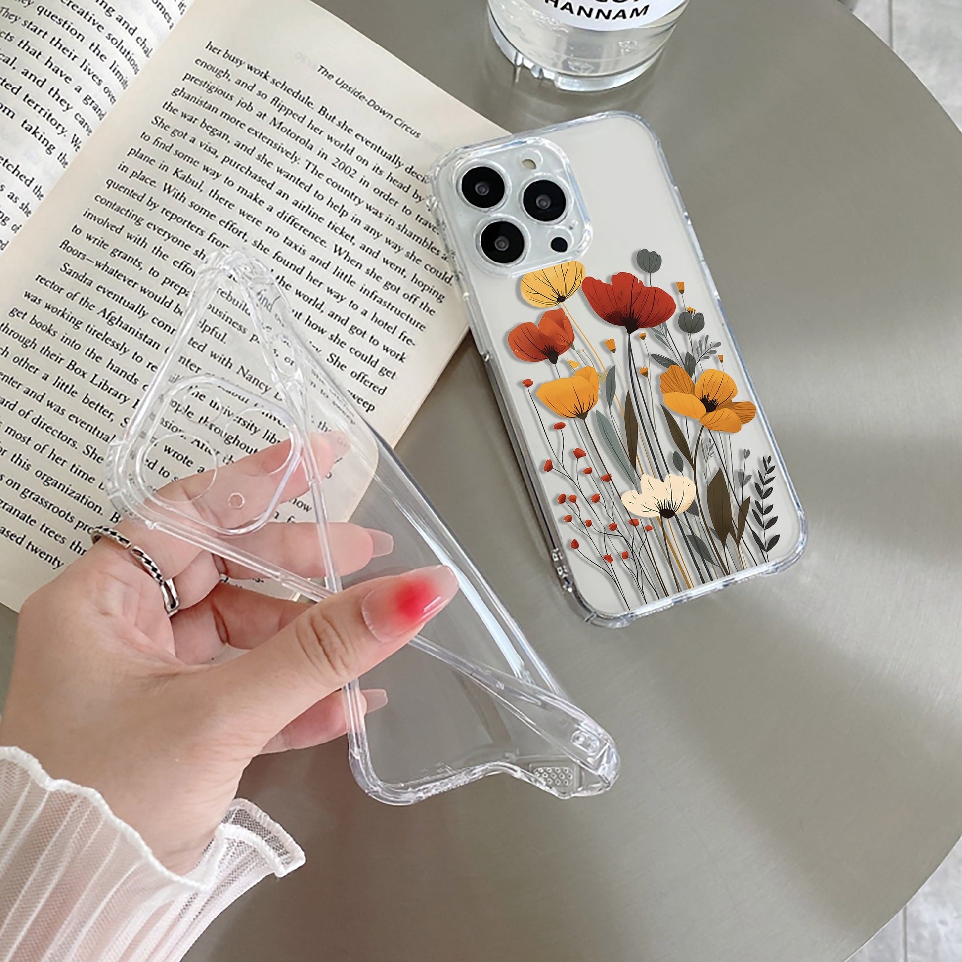 Clear phone case with floral design held by a hand, next to an open book and glass.